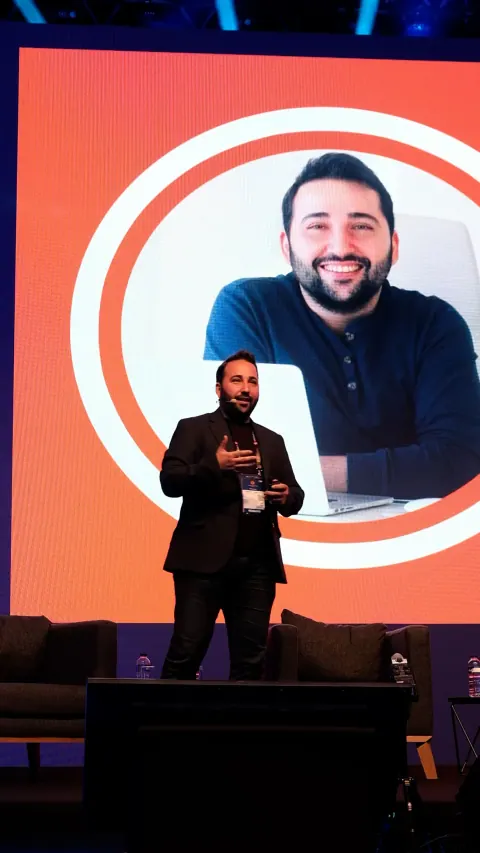 Kaan Gülten speaking on a stage with a large screen